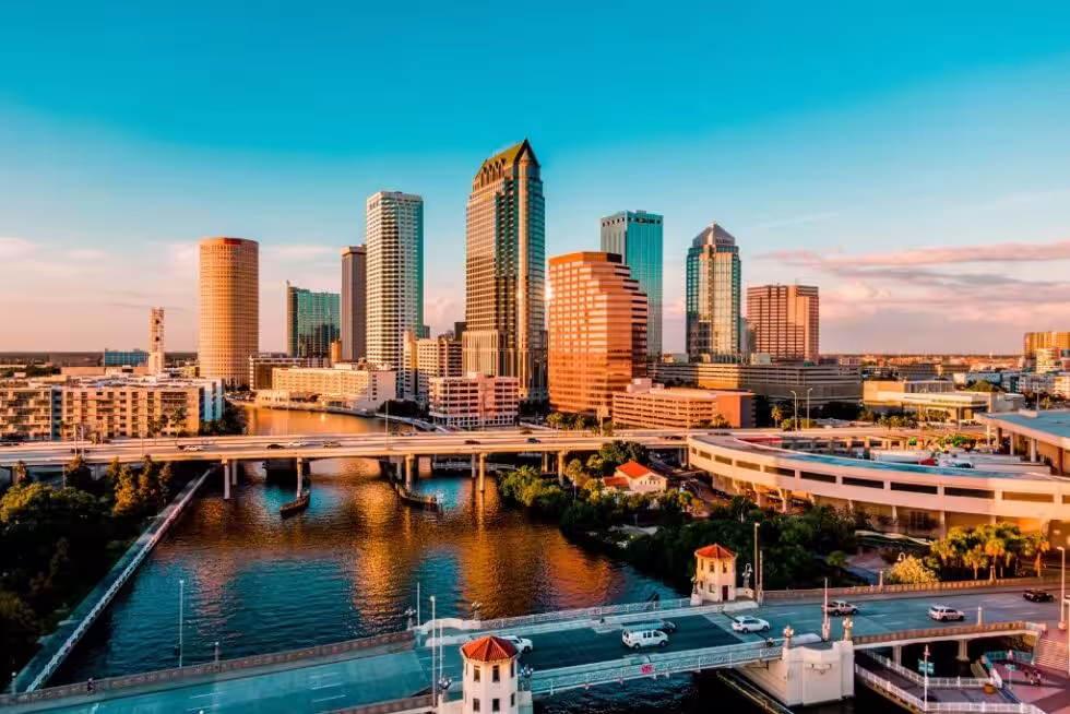 Tampa skyline beyond a river, bridges, and elevated roads in warm evening light.