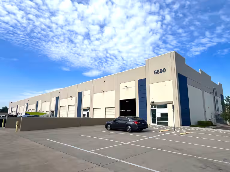 Commerce 270 in Commerce City, Colorado: building 5690 E 56th Avenue, a tan and blue tilt-up warehouse with grade-level doors, an open loading bay, a glass office entry, and striped parking under a partly cloudy sky